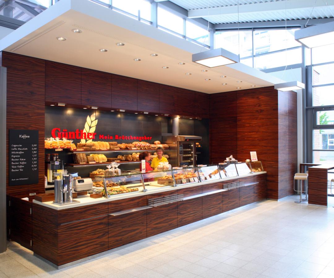 TTR-60, Foto: Christoph Edelhoff Verkaufstresen mit Rückraummöbel und Wandverkleidung aus dunkel furniertem Holz, Tresenaufsatz aus Glas und Edelstahl für eine Bäckerei